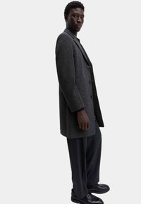 Cappotto di lana grigio con un design sartoriale, due bottoni e tasche laterali, abbinato a pantaloni a righe verticali scuri e scarpe nere.