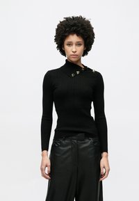 Schwarzer, gerippter Strickpullover mit hohem Kragen und dekorativen Knöpfen auf der Schulter, kombiniert mit schwarzen Lederhosen.