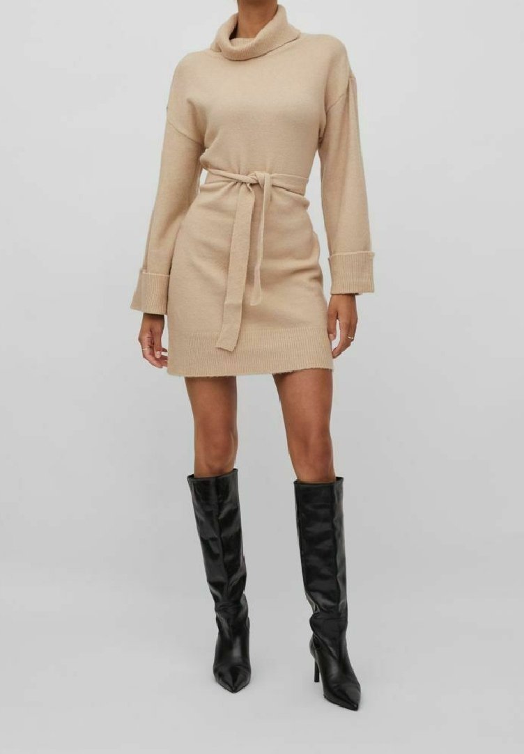 Femme portant une robe-pull beige en tricot avec une ceinture à la taille et des bottes noires montantes à talons pointus, debout devant un fond gris uni.