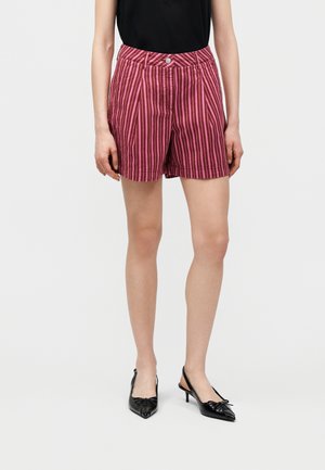 Frau trägt hoch taillierte Shorts mit pinken und dunkelroten vertikalen Streifen, ein schwarzes ärmelloses Oberteil und schwarze Slingback-Pumps mit Schleifen.