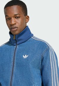 Jeansjacke mit hohem Kragen, blauer Farbe, Reißverschluss und weißem Logo. Verfügt über kontrastierende Streifen entlang der Schultern.