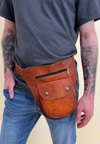 Tan leather fanny pack with a smooth texture, featuring a zippered pocket and a front snap-flap pocket. Worn around the waist.