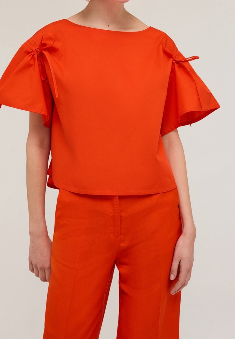 Femme portant un haut orange vif avec des manches courtes à volants nouées par de petits nœuds et un pantalon orange assorti sur un fond uni.