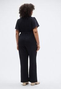 Fekete overál rövid ujjakkal, elasztikus derékkal és bő szárú kialakítással. A modell bézs magassarkút visel, texturált anyagból.