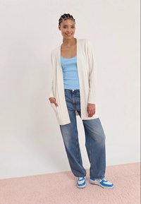 Young woman with braided hair wearing a white cardigan, blue tank top, wide-leg jeans, and blue-white sneakers, standing on pink carpet.