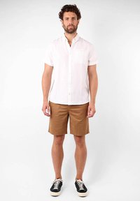 Chemise blanche à manches courtes, shorts marron, baskets noires avec semelles blanches. Le modèle se tient contre un fond clair, les bras détendus.