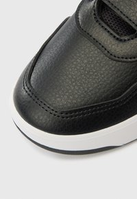 Mustast nahast toss, millel on tekstuuriga pind, sees on Velcro rihm, valge talla ja õmbluse detailid, esitades madala profiiliga disaini.