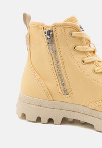 Botă galbenă tip high-top, fabricată din pânză texturată, cu fermoar pe lateral și talpă de cauciuc bej, dotată cu crampoane adânci pentru tracțiune.