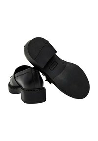 Mocassini neri in pelle con tacco spesso, suola in gomma testurizzata e dettaglio con cinturino, una scarpa mostra la suola inferiore con il nome del marchio.