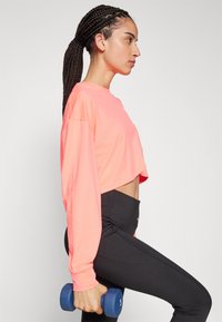 Neon rosa cropped langermet skjorte, kombinert med høyde-justerte svarte leggings, holder en blå manual med nummer 3 synlig.