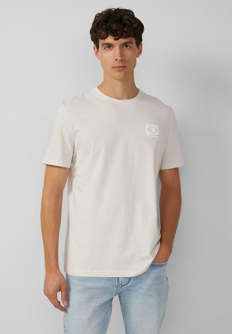 Lichtbeige katoenen t-shirt met korte mouwen, ronde hals en een klein wit grafisch logo op de linkerborst. Casual pasvorm.
