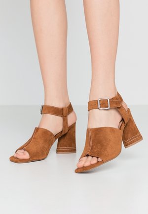 Bruine suède sandalen met blokhak, open teen, enkelbandje en zilveren gesp, gedragen aan de voeten tegen een effen achtergrond.