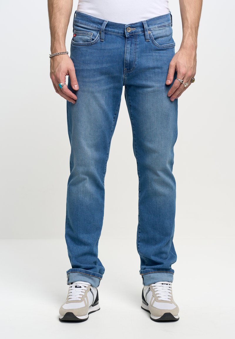 Vaqueros de mezclilla azul claro con un corte recto, que presentan un diseño clásico de cinco bolsillos y costuras visibles. Combinados con zapatillas grises y blancas.
