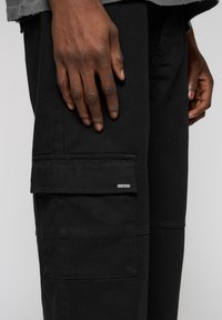 Nahaufnahme einer dunkelhäutigen Hand, die auf einer schwarzen Hose mit einer Cargotasche ruht, die ein kleines rechteckiges Metallschild aufweist.