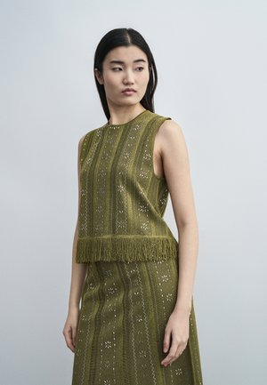 Γυναίκα με μακριά μαύρα μαλλιά φοράει αμάνικη τοπ ελιάς με μοτίβο και κρόσια και ταιριαστή φούστα, κοιτώντας ελαφρώς στο πλάι.