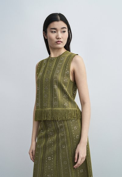 Femme aux longs cheveux noirs portant un haut sans manches vert olive à motifs avec des franges et une jupe assortie, regardant légèrement sur le côté.
