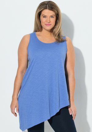 Frau mit schulterlangem Haar, die ein ärmelloses blaues asymmetrisches Top und schwarze Leggings trägt und vor einer weißen Wand steht.