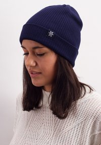 Navy ribbed knit beanie with a turned-up edge and a small black logo patch, worn by a person with long dark hair.