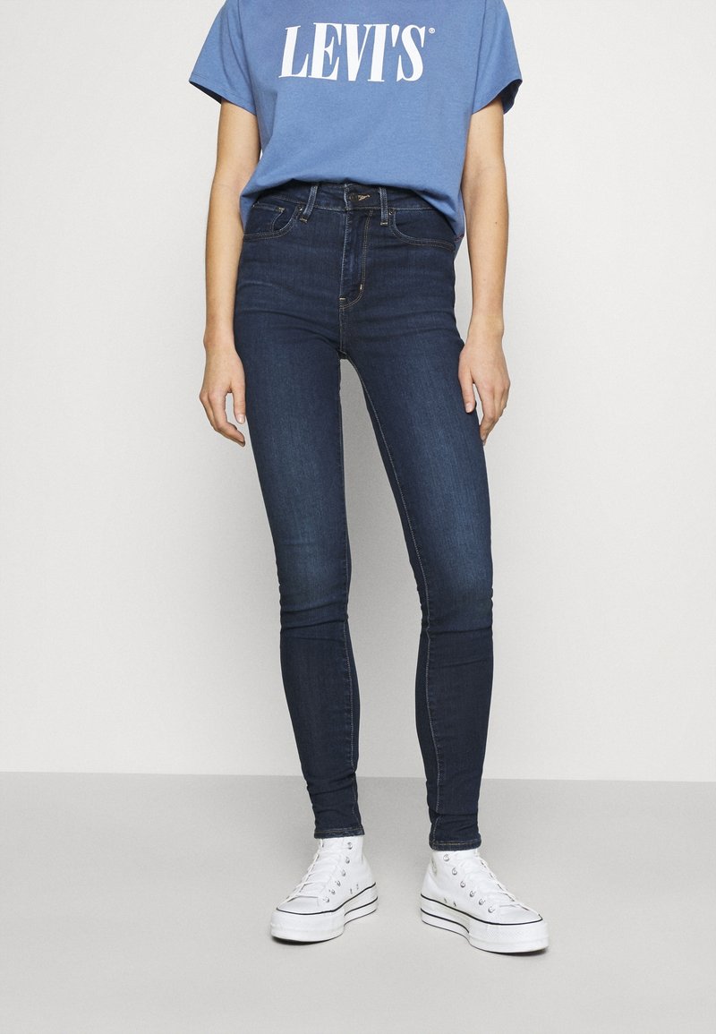 Jean skinny bleu foncé en denim, avec une taille haute et une coupe slim. Associé à des baskets montantes blanches et un t-shirt graphique bleu.