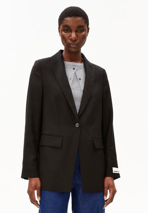 Woman wearing a tailored black blazer over a light gray top and blue jeans, standing against a plain white background.