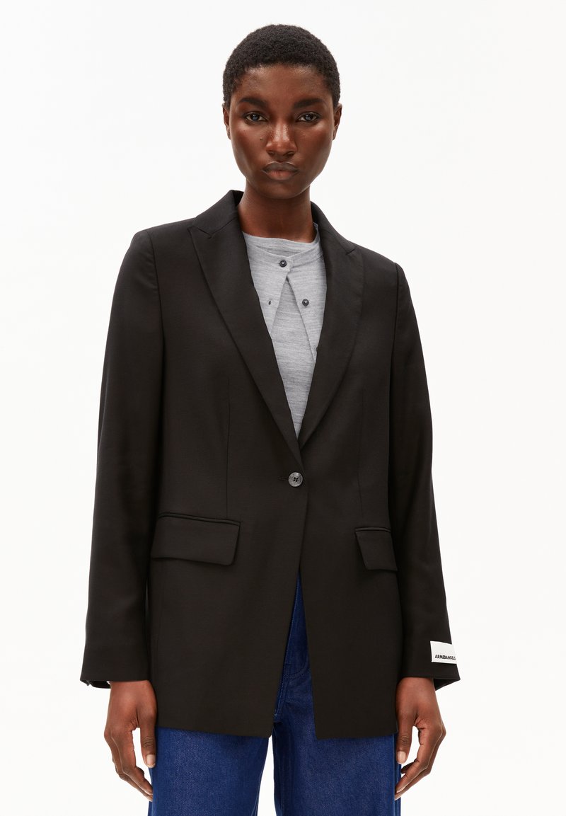 Vrouw in een op maat gemaakte zwarte blazer over een lichtgrijze top en blauwe spijkerbroek, staande tegen een effen witte achtergrond.