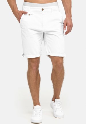 Mann trägt hochgekrempelte weiße Shorts mit braunen Knöpfen und weiße Sneakers, steht mit einer Hand in der Tasche vor neutralem Hintergrund.