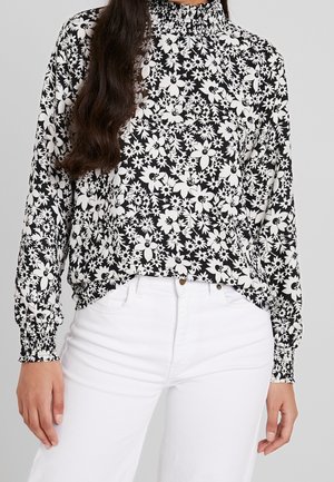 Czarna i biała bluzka w kwiatowy wzór z długim rękawem, marszczonymi mankietami i wysokim golfem, zestawiona z białymi, wysokim stanem spodniami.