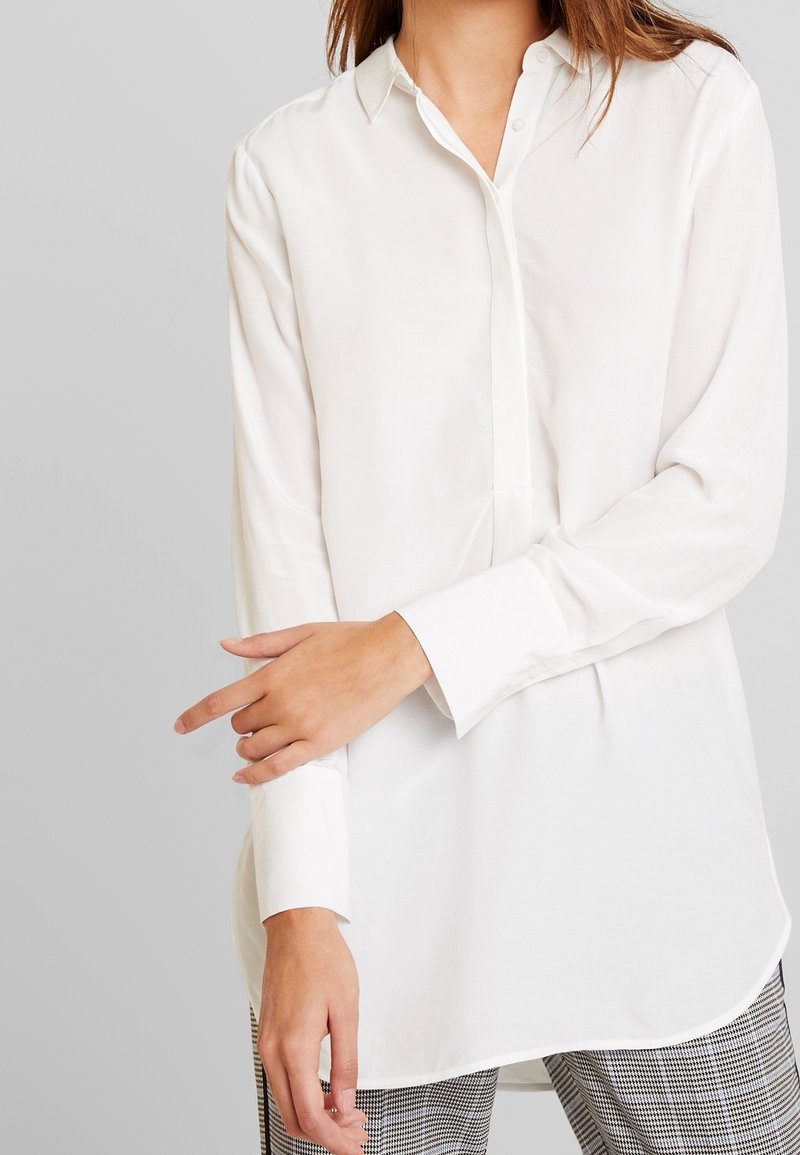 Blouse blanche, manches longues, boutonnage sur le devant, tissu doux, ourlet courbé, texture légère, portée avec un pantalon à carreaux.