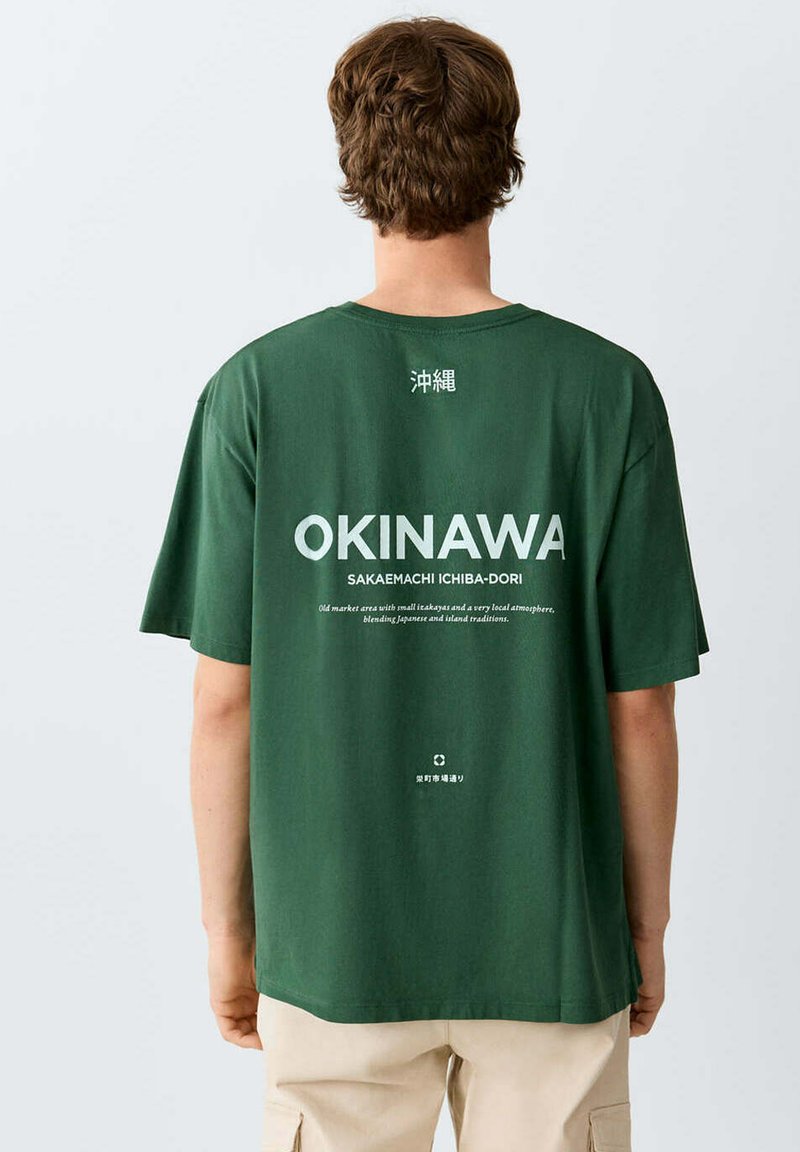 Homme portant un T-shirt vert avec texte blanc Okinawa et caractères japonais, debout de dos, pantalon beige visible de la taille vers le bas.