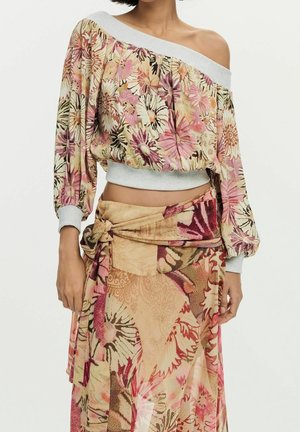 Donna che indossa un top floreale scollato con maniche lunghe e una gonna a portafoglio floreale abbinata, legata in vita in tonalità rosa e beige.