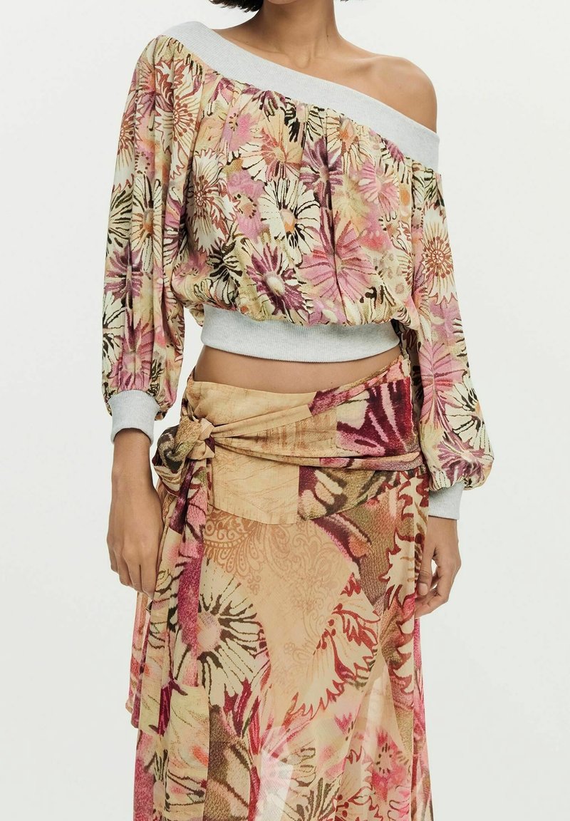 Femme portant un top floral à épaules dénudées avec manches longues et une jupe portefeuille assortie nouée à la taille dans des tons roses et beiges.