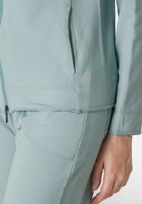 Giacca a zip color turchese chiaro con tasche, dalla texture morbida e dettagli di cucitura in evidenza. Presenta un logo dal design circolare sull'orlo.