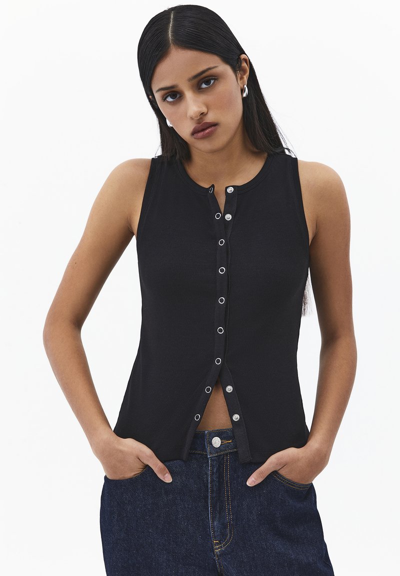 Jeune femme aux longs cheveux foncés porte un top noir sans manches boutonné et un jean bleu, debout les mains dans les poches devant un fond blanc.