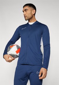 Marineblauwe sportjack met rits en lange mouwen, witte zijaccenten en Nike-logo. Model houdt een voetbal vast met zwarte en rode details.