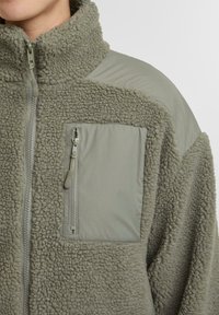 Grau Fleece-Jacke mit hohem Kragen, Druckknopf-Reißverschluss und glattem Stoffpanel auf den Schultern. Verfügt über eine zipfähige Brusttasche und strukturierte Oberfläche.