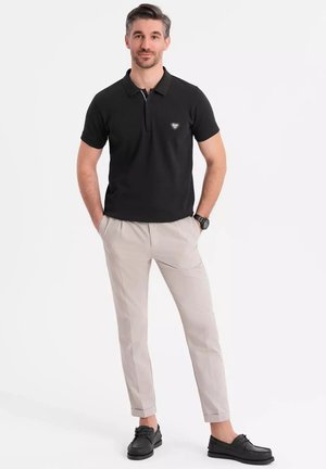 Sort polo skjorte med krave, grå logo på brystet, parret med lyse beige bukser og sorte sko. Stående mod en hvid baggrund.
