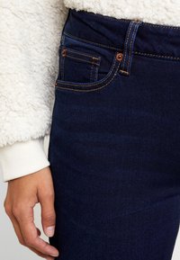 Jeans de mezclilla azul oscuro con costuras naranjas en contraste, que presentan un detalle en el bolsillo frontal. La tela tiene una apariencia suave y ligeramente texturada.