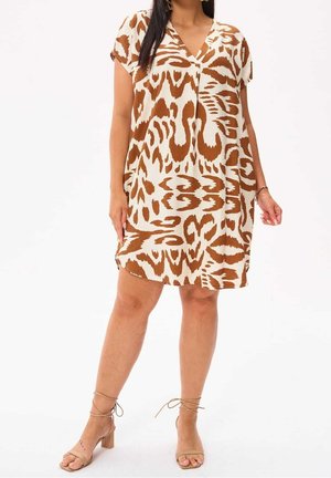 Kvinde iført en løs, knælængde, kortærmet kjole med brunt og hvidt abstrakt dyreprint og beige sandaler med strop og blokhæl.