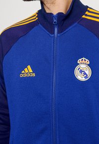 Blå jacka med dragkedja och kontrasterande mörkblå raglanärmar, med gul adidas-logotyp och Real Madrid-vapen. Slät, atletisk materia.