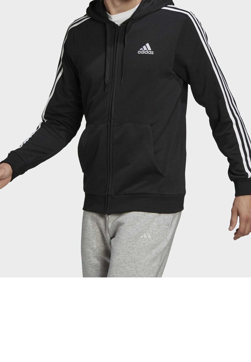 Uomo che indossa una felpa nera Adidas con zip e strisce bianche sulle maniche, insieme a pantaloni grigi Adidas, su uno sfondo neutro.