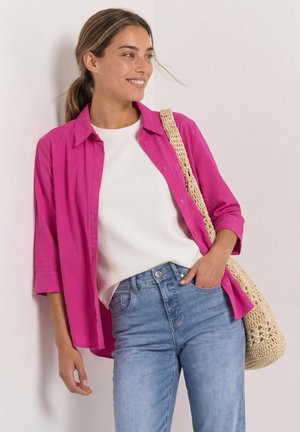 Jonge vrouw met paardenstaart die een roze shirt draagt over een witte top en blauwe spijkerbroek, met een beige gehaakte tas op haar schouder.