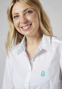 Chemise blanche à boutons avec un blason brodé bleu et un col à motif floral, fabriquée dans un tissu lisse au toucher croquant.