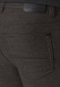 Dunkelgraue Jeanshose mit strukturierter Oberfläche, versehen mit einem Lederetikett am Bund und einer einzelnen Gesäßtasche.