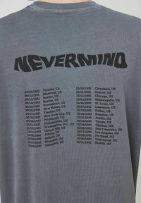 T-shirt en coton gris présentant le texte noir "NEVERMIND" dans le haut du dos ; liste les dates et lieux de concerts sous forme de colonne en dessous.