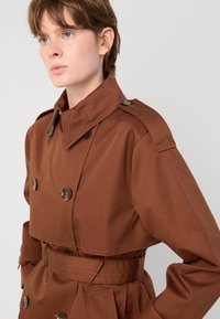 Brązowy płaszcz trench z gładkiego materiału, z podwójnym zapięciem na guzik, dużymi guzikami i paskiem w talii dla podkreślenia sylwetki.