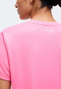 Γυναίκα που φοράει ροζ μπλουζάκι Under Armour, εμφανιζόμενη από πίσω, με λογότυπο κοντά στον λαιμό και μαλλιά δεμένα σε κότσο.