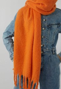 Personne portant une écharpe orange vif à poils avec des franges sur une veste en denim bleue boutonnée et un jean assorti, les mains posées sur les hanches.