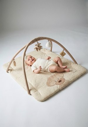 Baby lying on beige play mat with bear design, under soft toy arch holding plush toys in neutral-toned room with white background.
