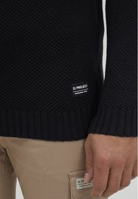 Dunkelblaues gestricktes Sweatshirt mit Texturmuster, gerippten Bündchen und einem Markenpatch mit der Aufschrift "11 PROJECT" an der unteren Seite.