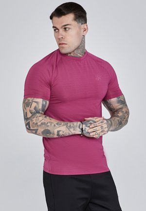 Magenta aansluitend shirt met korte mouwen en een textuurdesign, geribbelde halslijn en subtiele logo-accent. Model poseert met gekruiste armen.